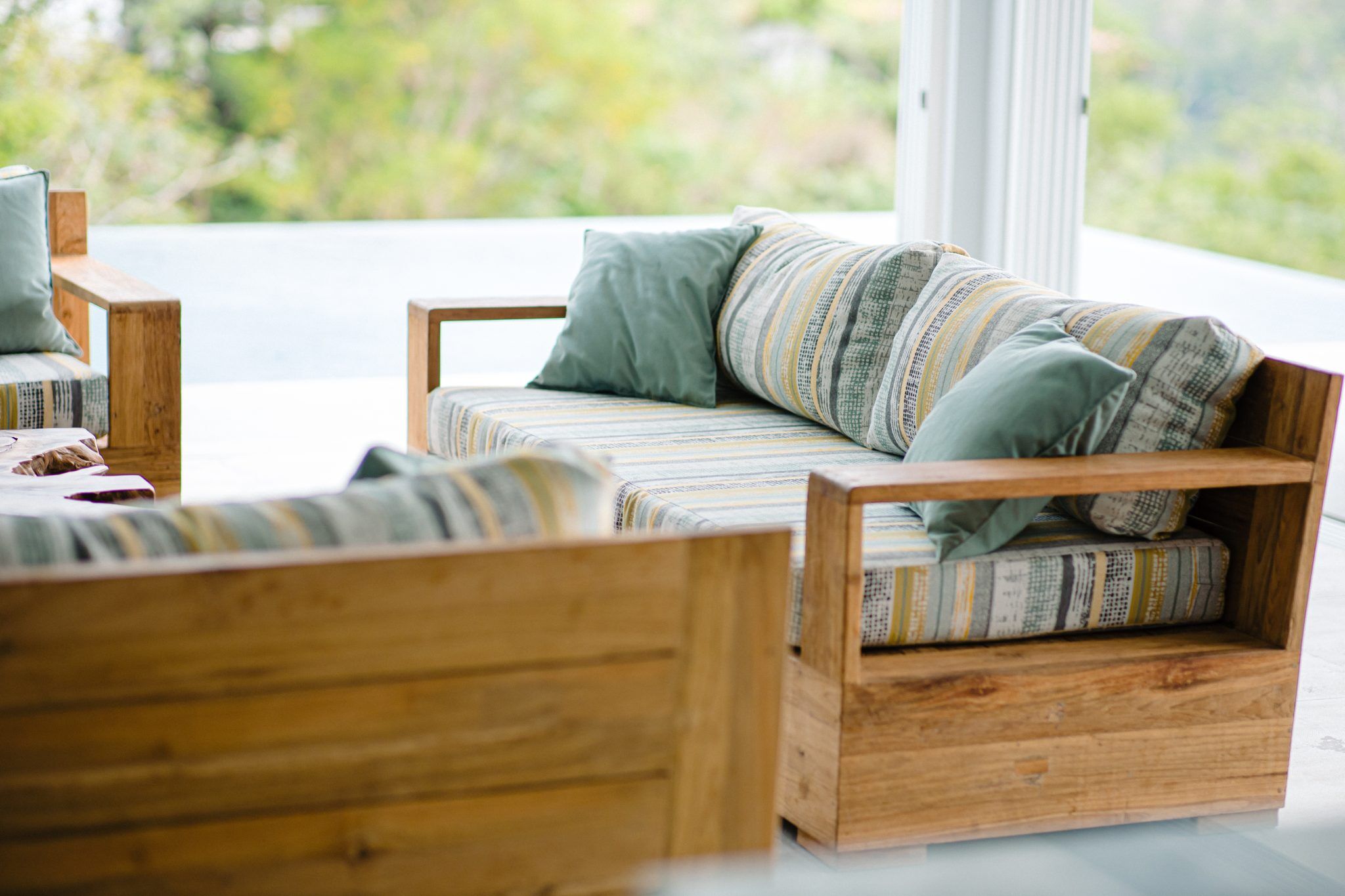 Wooden patio furniture with striped cushions and green pillows on a terrace overlooking a blurred natural background.
